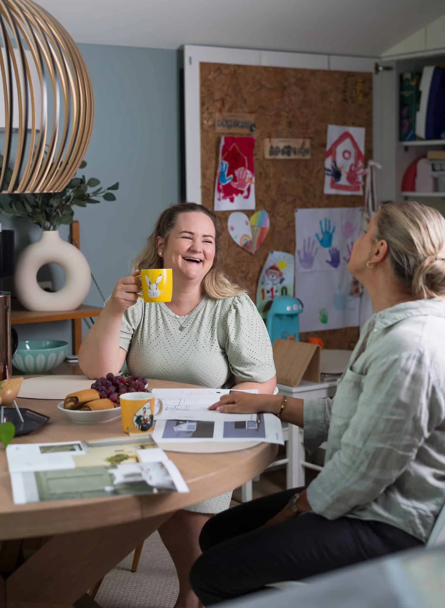 To kvinner som ler og prater over kaffe i et koselig hjemmekjøkken.