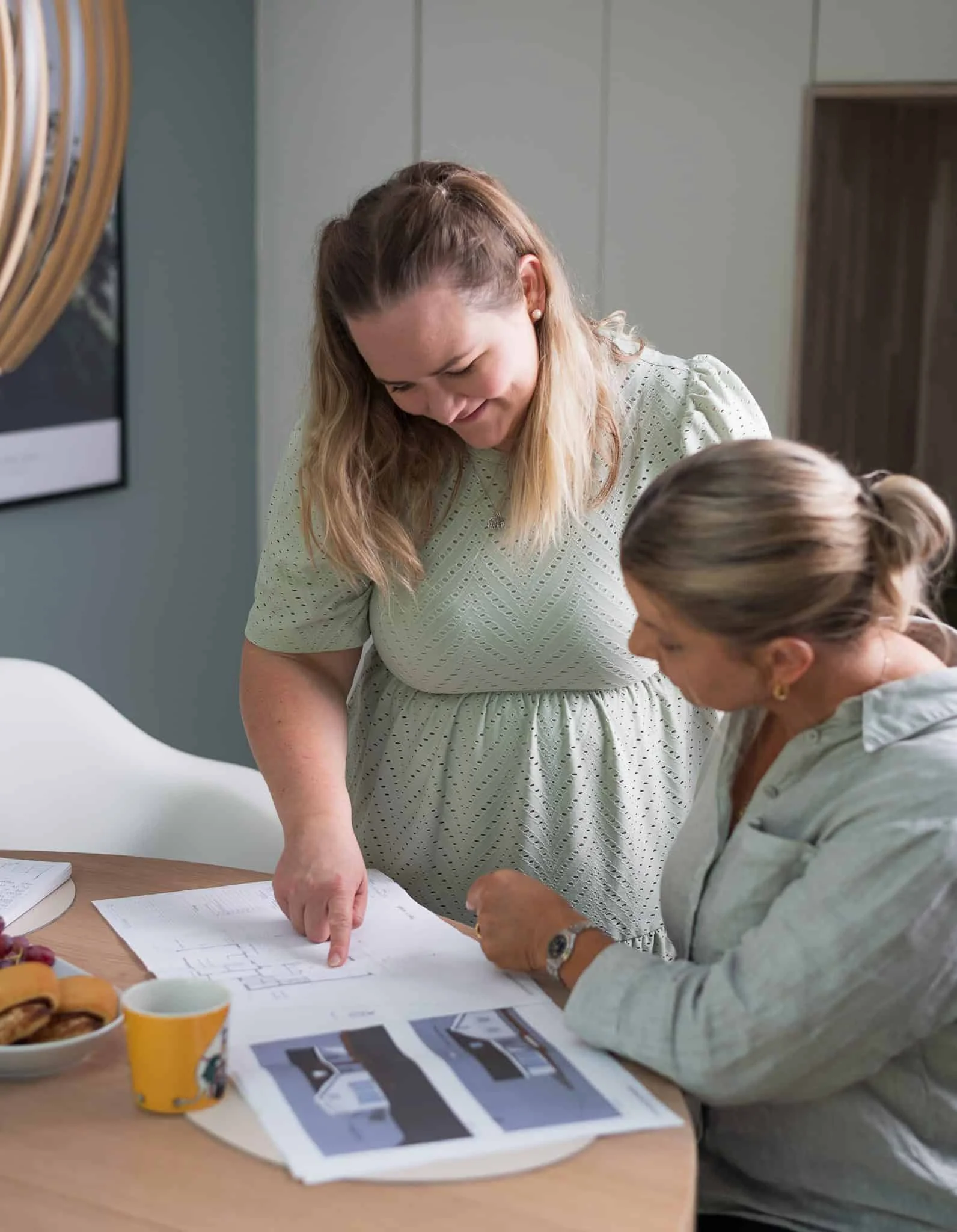 To kvinner diskuterer design av minnesmerke, med bilder og skisser på bordet.