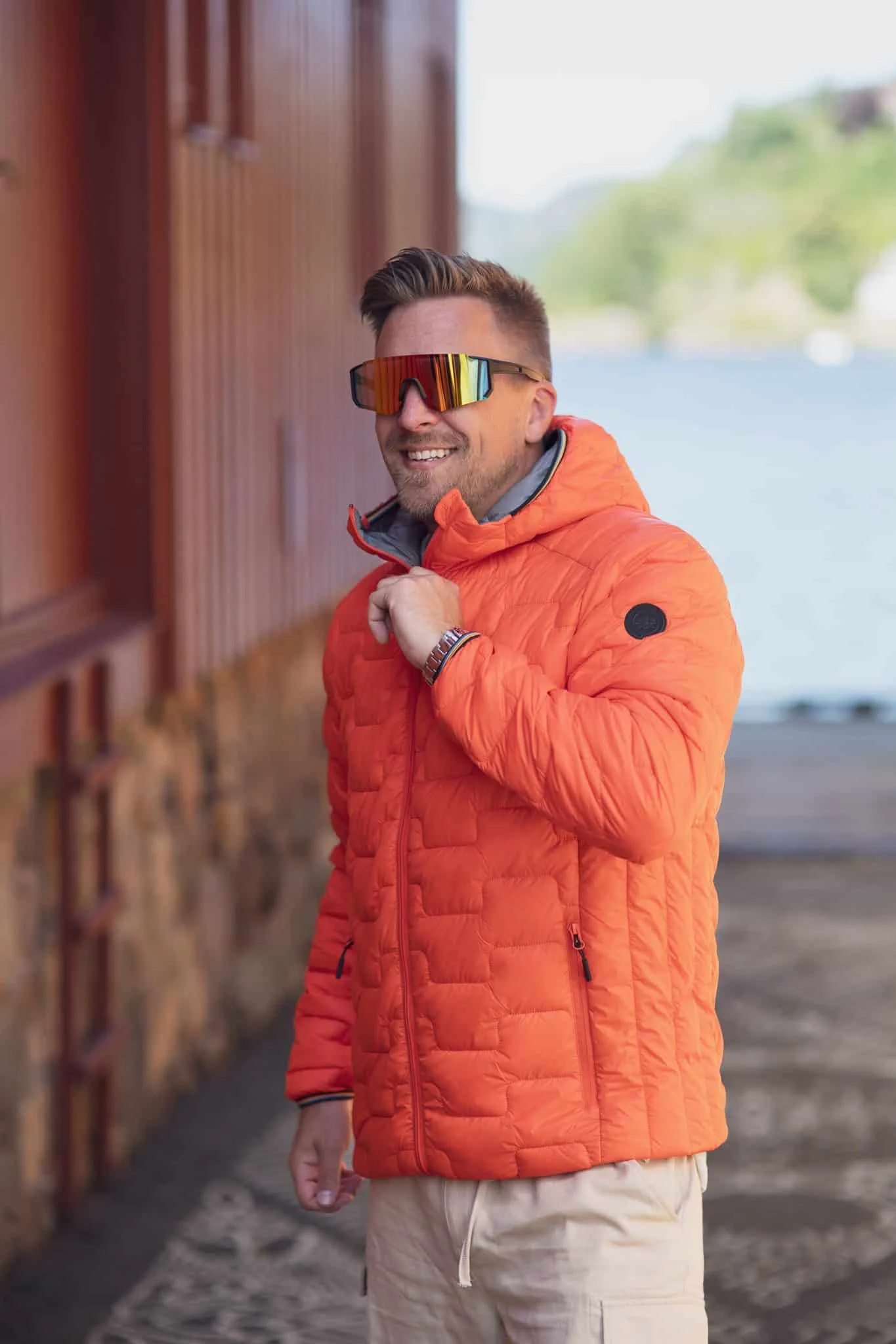 Portrett av en mann med solbriller og oransje jakke, smilende og glad, foran en vannkyst i naturlig.