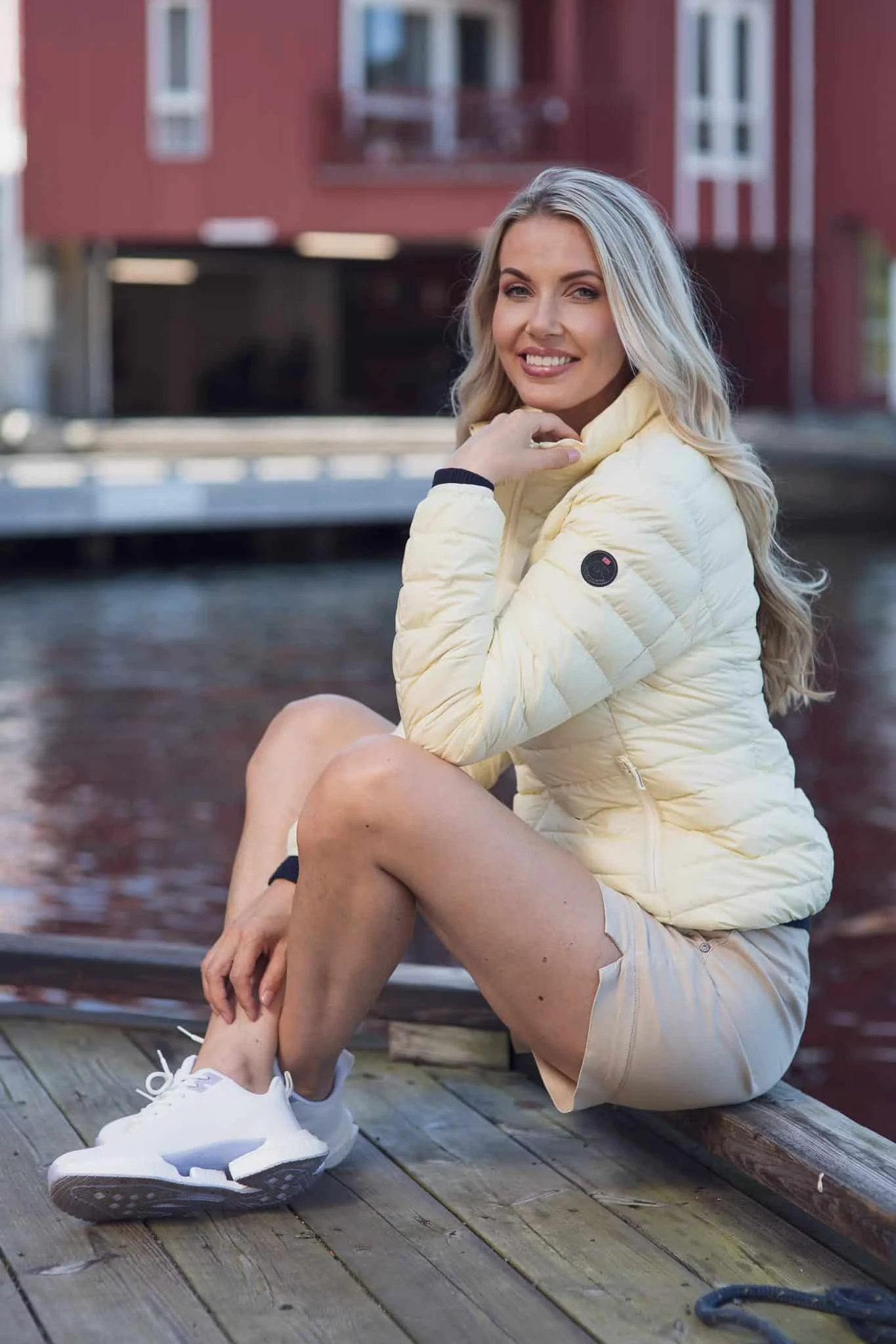 Smilende kvinne i lys jakke ved vannet, sittende på brygge, med moderne bybakgrunn.