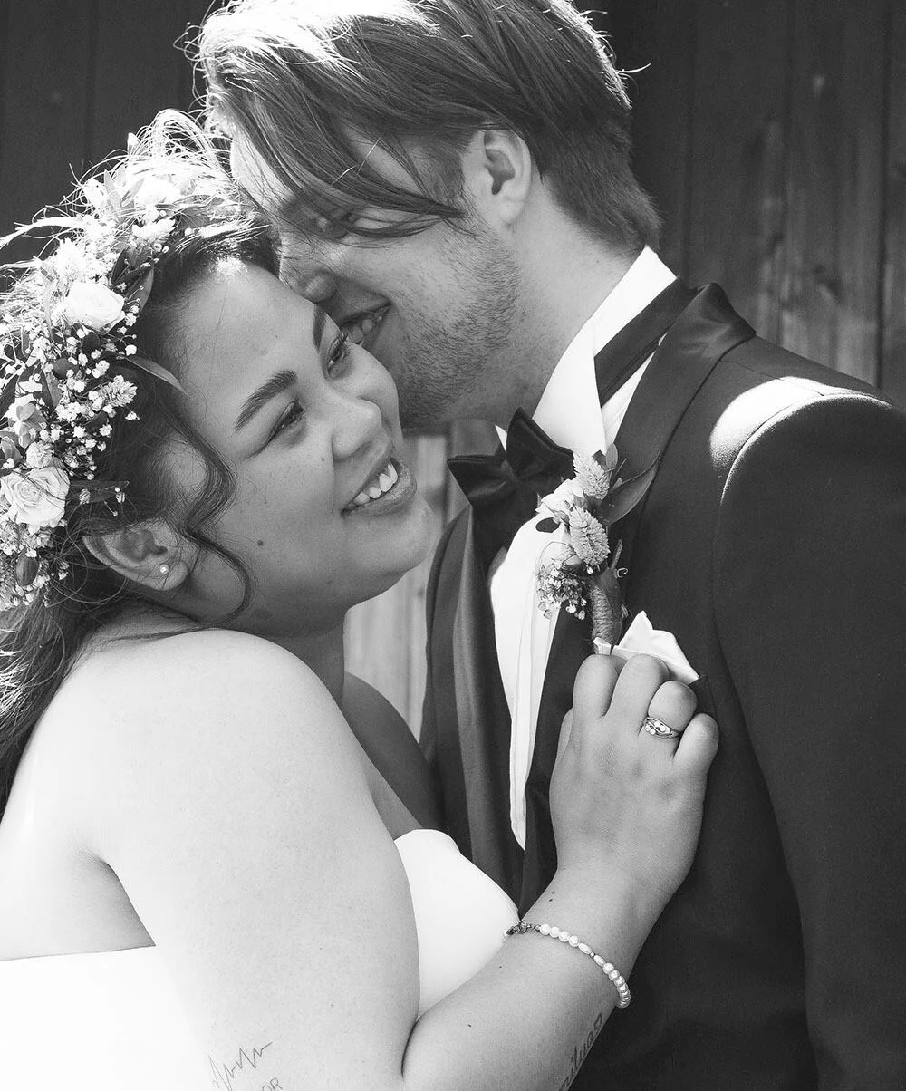 Brud med blomsterkrans smiler til brudgom i dress som hvisker henne i øret, fotografert i en tidløs, romantisk stil.