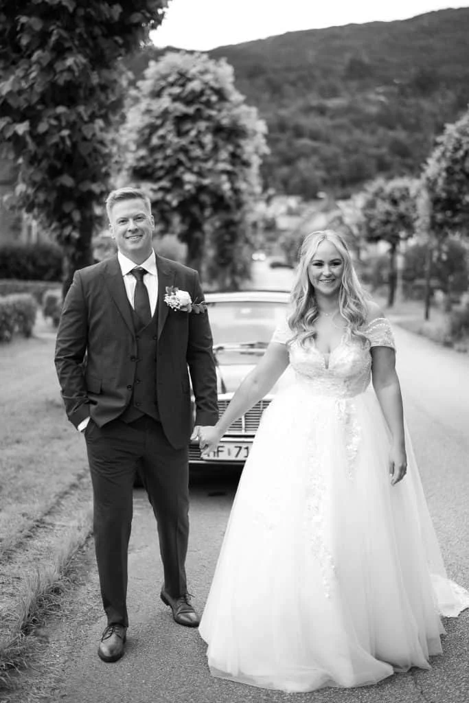 Brudepar som smiler og holder hender foran en klassisk bryllupsbil, fotografert i svart-hvitt av Evig Minne