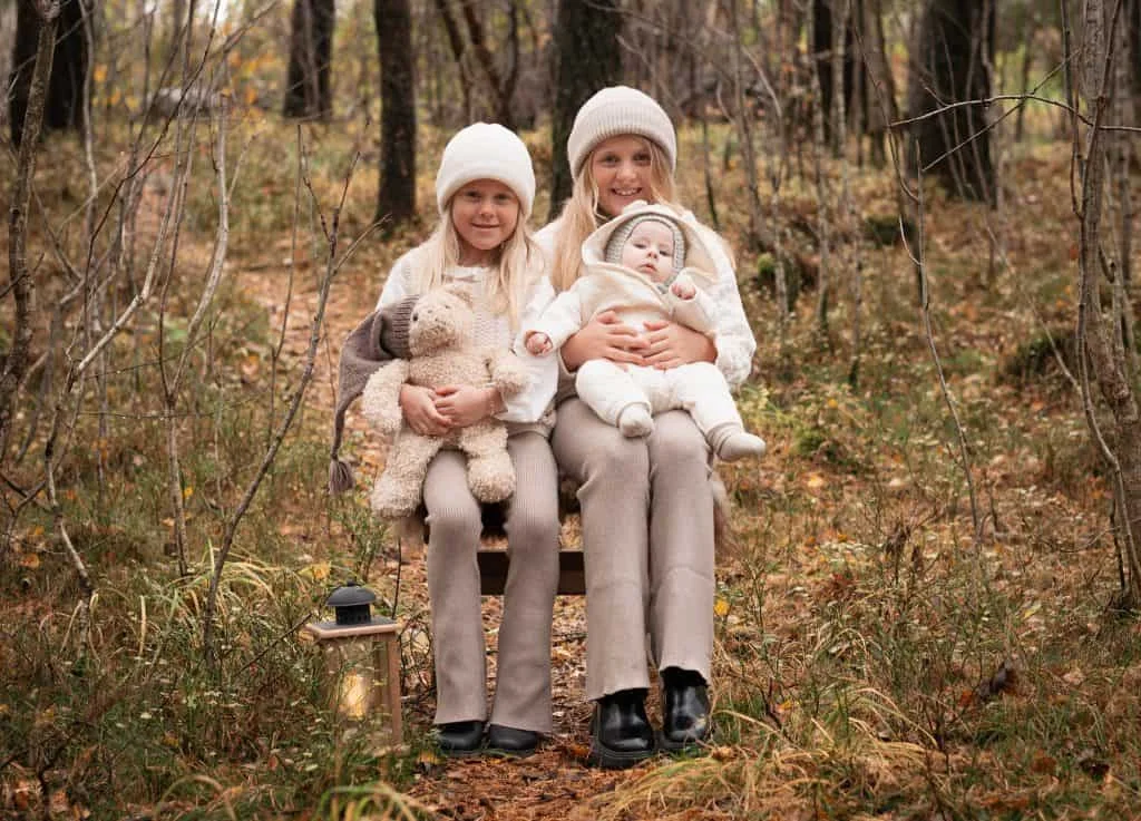 Søstre og en baby i beige strikkeklær og luer sitter på en benk i skogen under profesjonell nyfødtfotografering.
