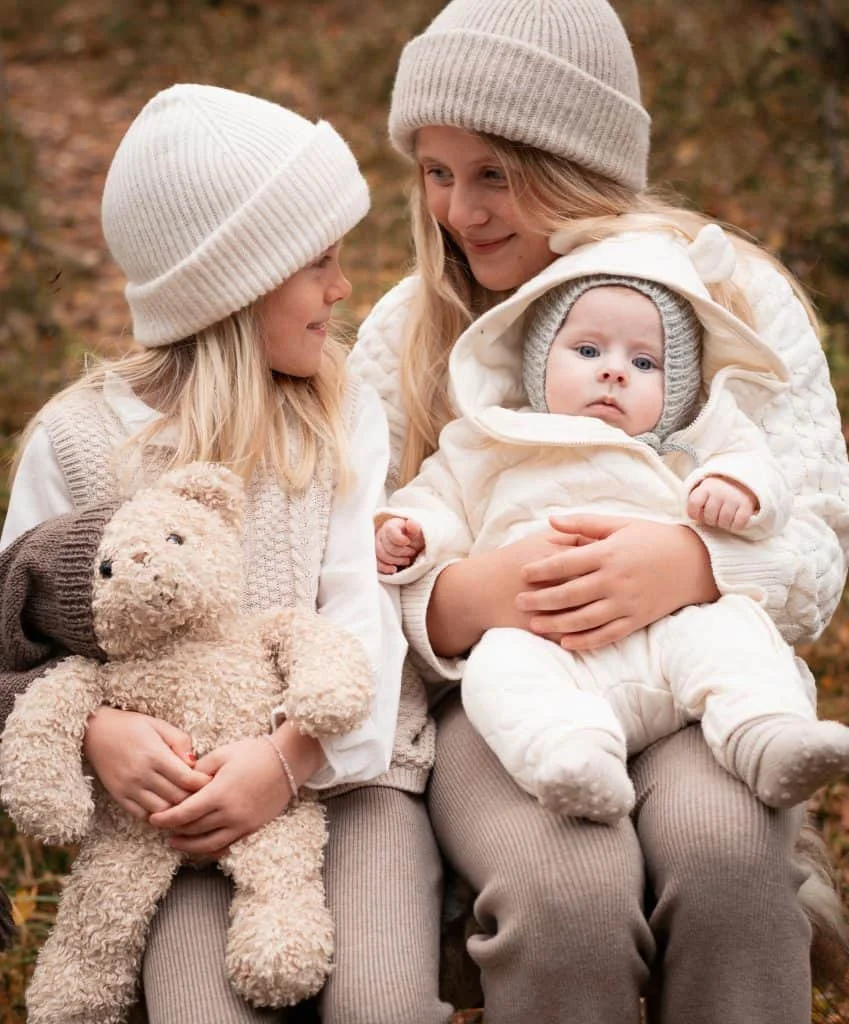 To eldre søstre holder en liten baby i lys dress ute i naturen under profesjonell nyfødtfotografering.