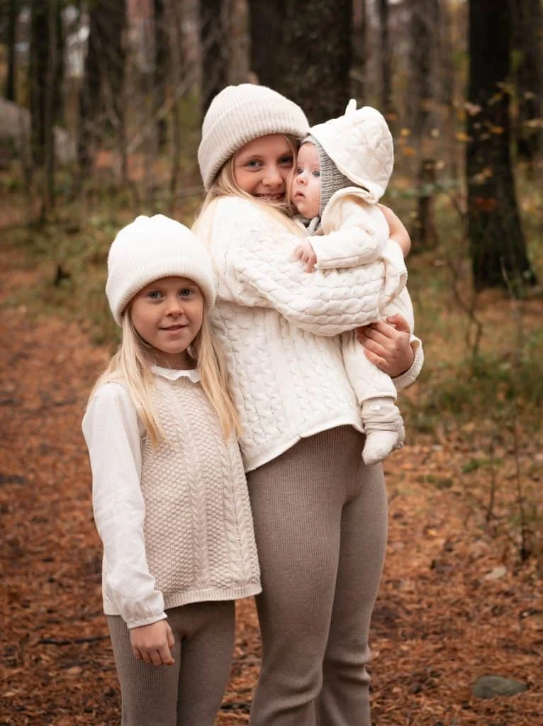 Tre søsken kledd i beige strikkeklær i en høstskog, der storesøster bærer på en baby under profesjonell nyfødtfotografering utendørs.
