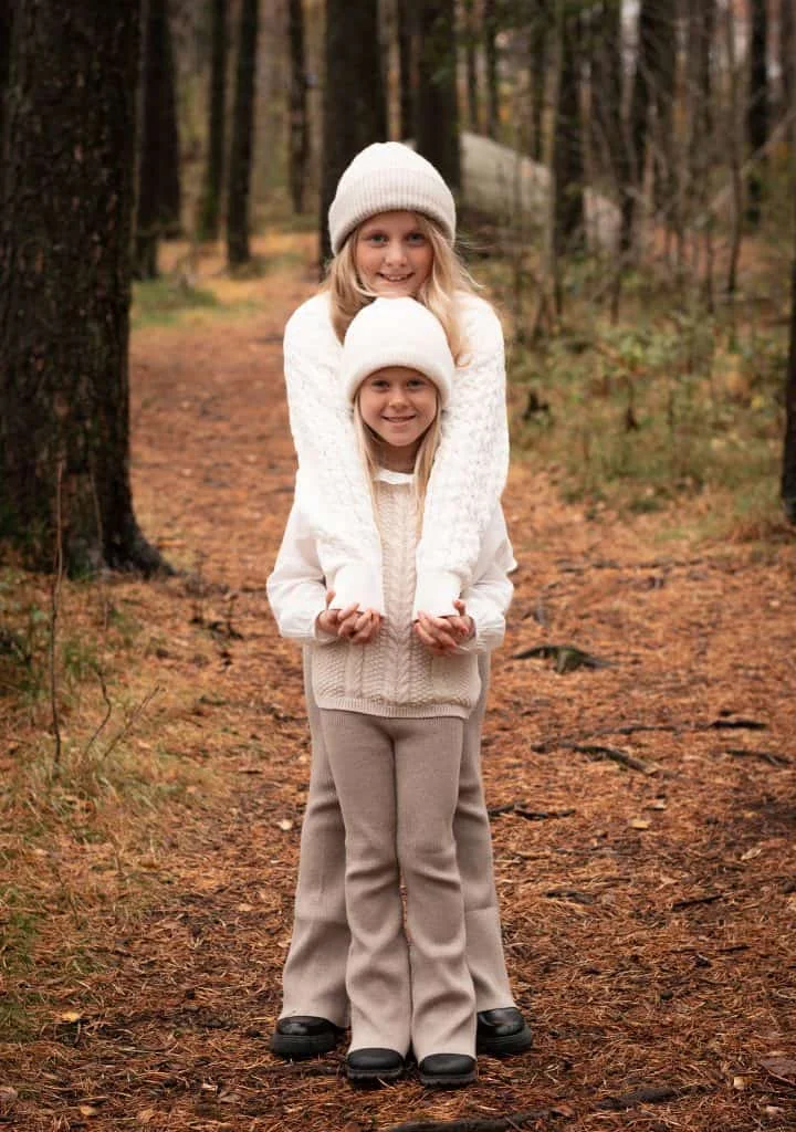 To smilende søstre i beige strikkegensere og luer står samlet i en høstskog, et stemningsfullt eksempel på familie- og barnefotografering.