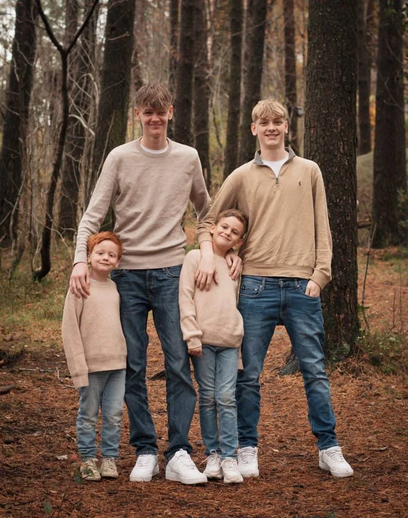 Fire brødre i ulike aldre står samlet i en skog under utendørs familie- og barnefotografering, kledd i matchende beige gensere.