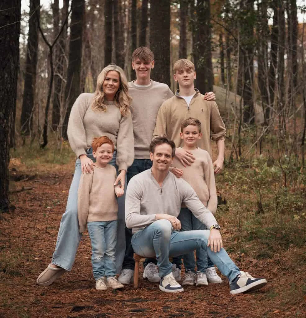 Smilende familie på seks kledd i beige gensere og jeans poserer sammen i en høstlig skog for profesjonell fotografering.