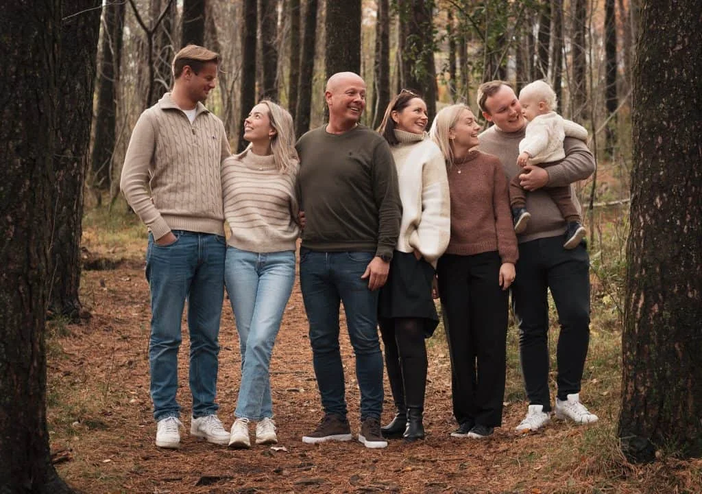 Storfamilie kledd i lune strikkegensere smiler til hverandre i en skog, perfekt for utendørs familie- og nyfødtfotografering.