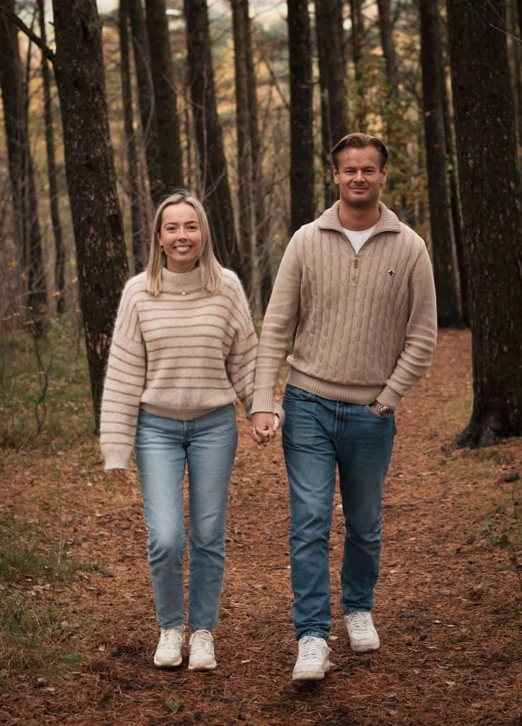 Et lykkelig par går hånd i hånd gjennom en høstskog i koordinerte beige gensere og blå jeans under familie- og parfotografering.