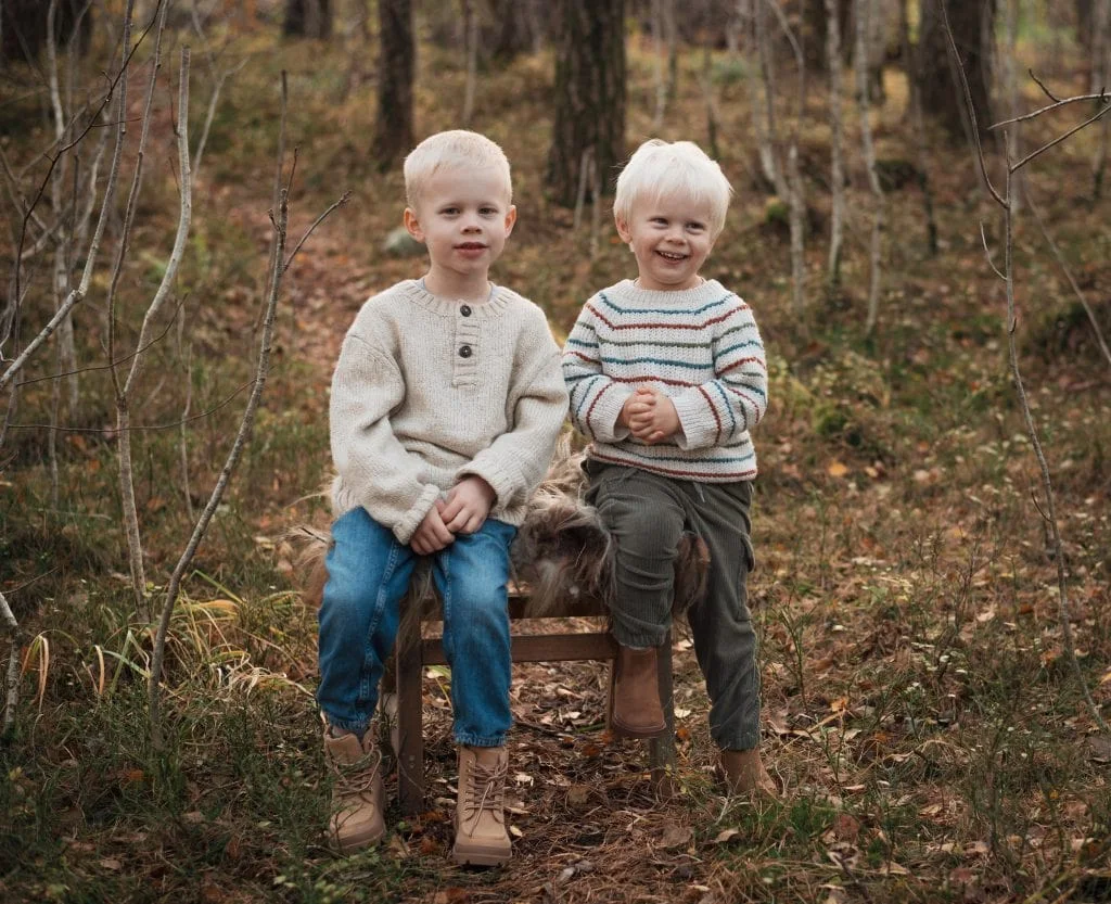To lyse gutter i strikkegensere sitter på en trebenk i skogen, et eksempel på naturlig barne- og familieportrettsfotografering.