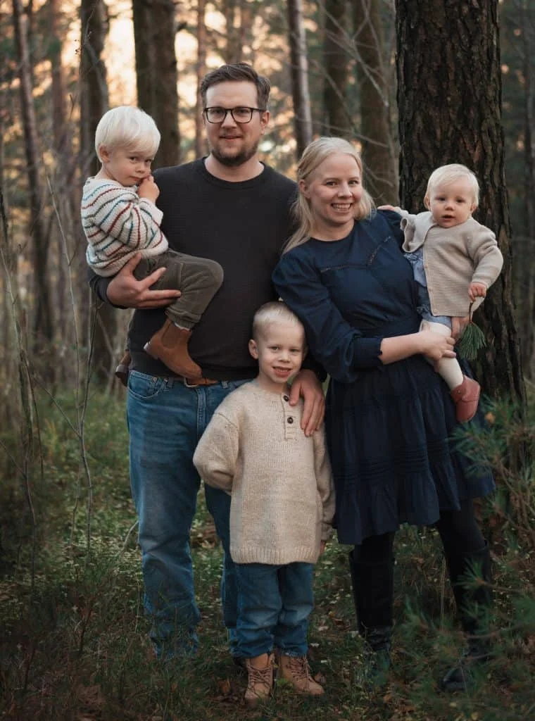 Småbarnsfamilie på fem smiler til kamera under utendørs familiefotografering i en lun skog med naturlig lys.