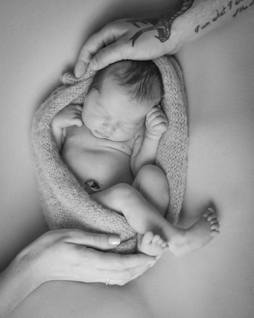 En sovende nyfødt baby er svøpt i et mykt strikket teppe og holdes varsomt av foreldrenes hender i et fotostudio.