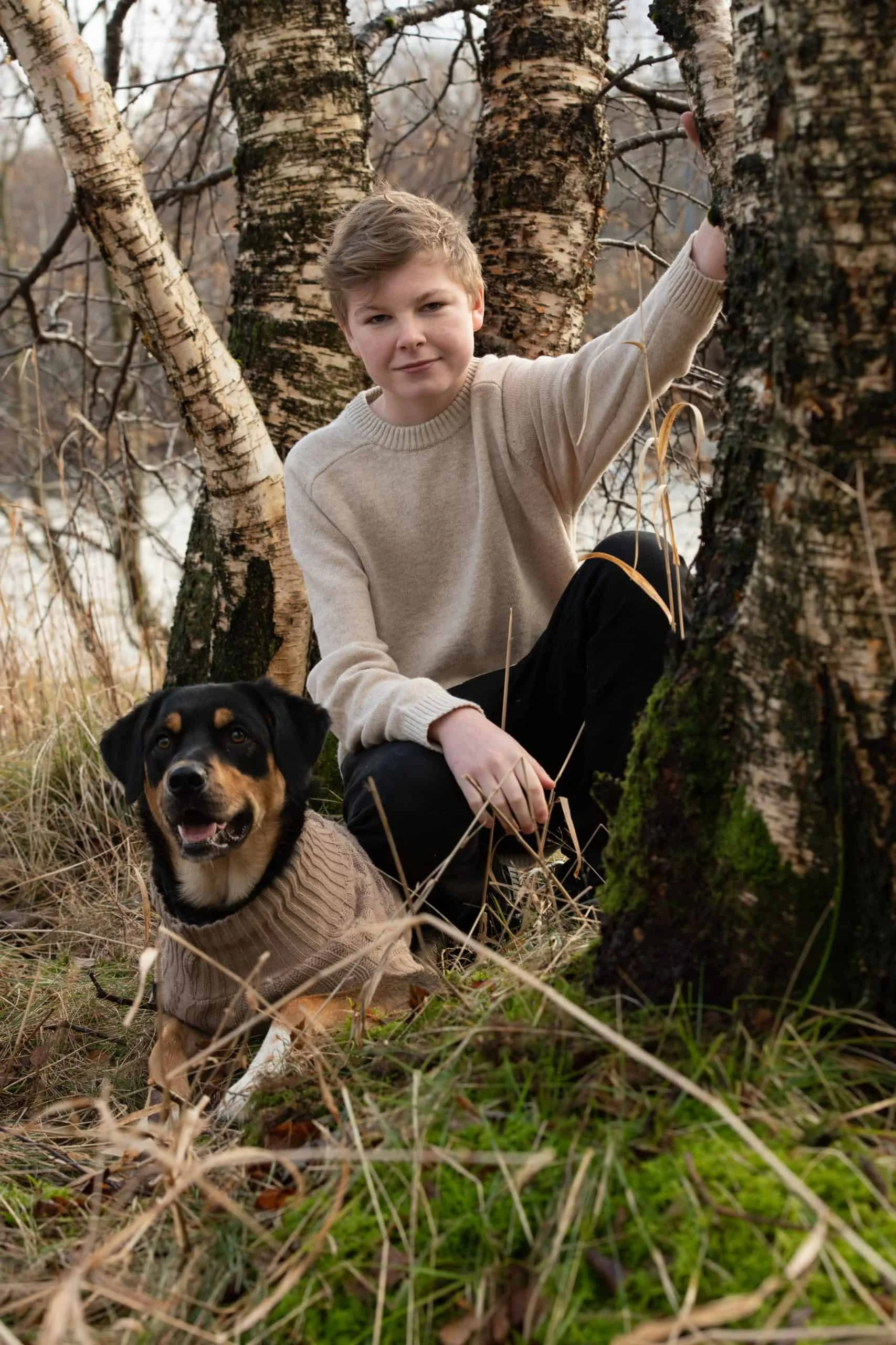 En ung gutt i beige genser sitter i skogen ved siden av en hund ikledd en matchende strikkegenser under profesjonell familie- og barnefotografering.