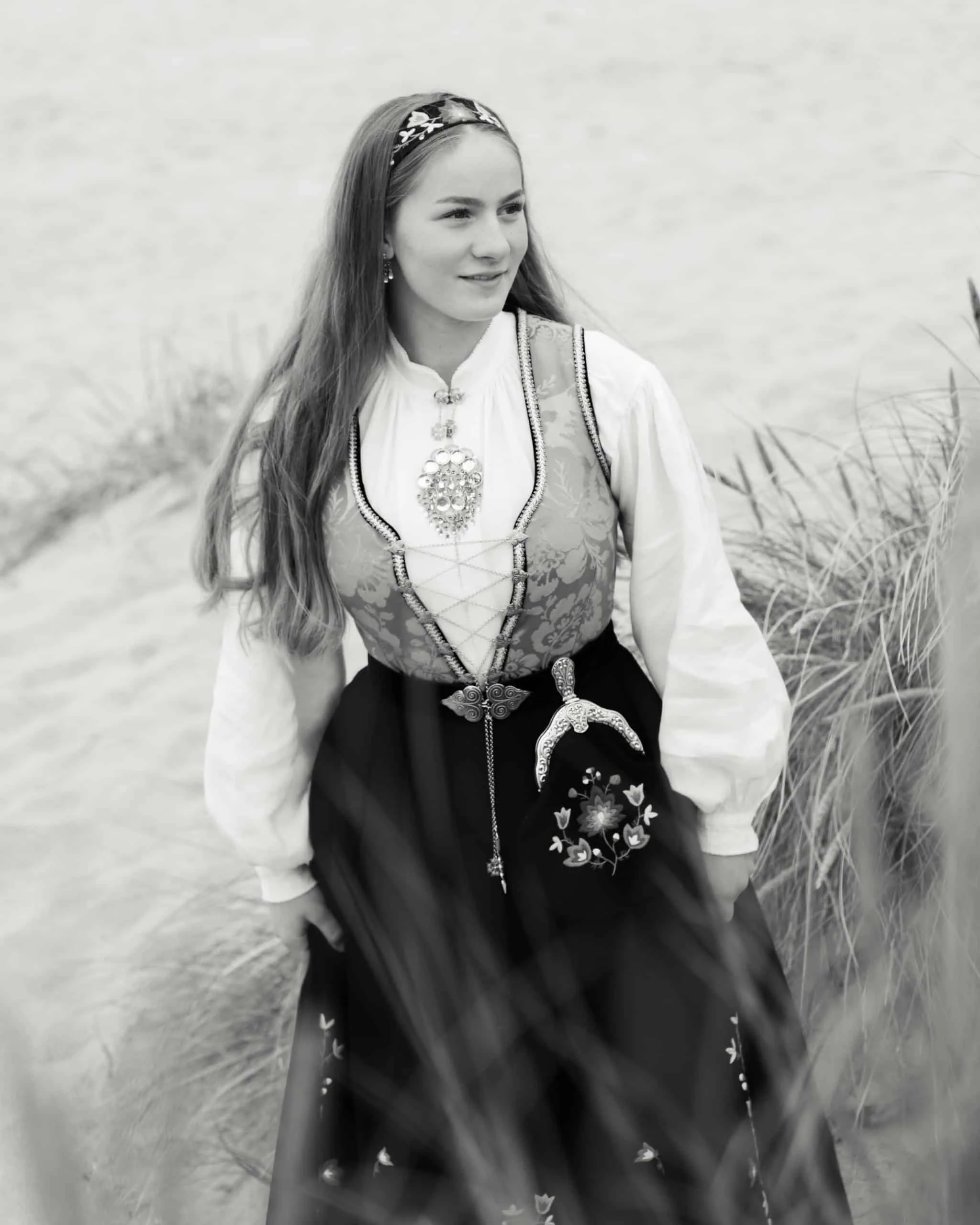 Ung kvinne kledd i tradisjonell norsk bunad med sølje og broderier, fotografert i et naturlig utemiljø med strandgress.