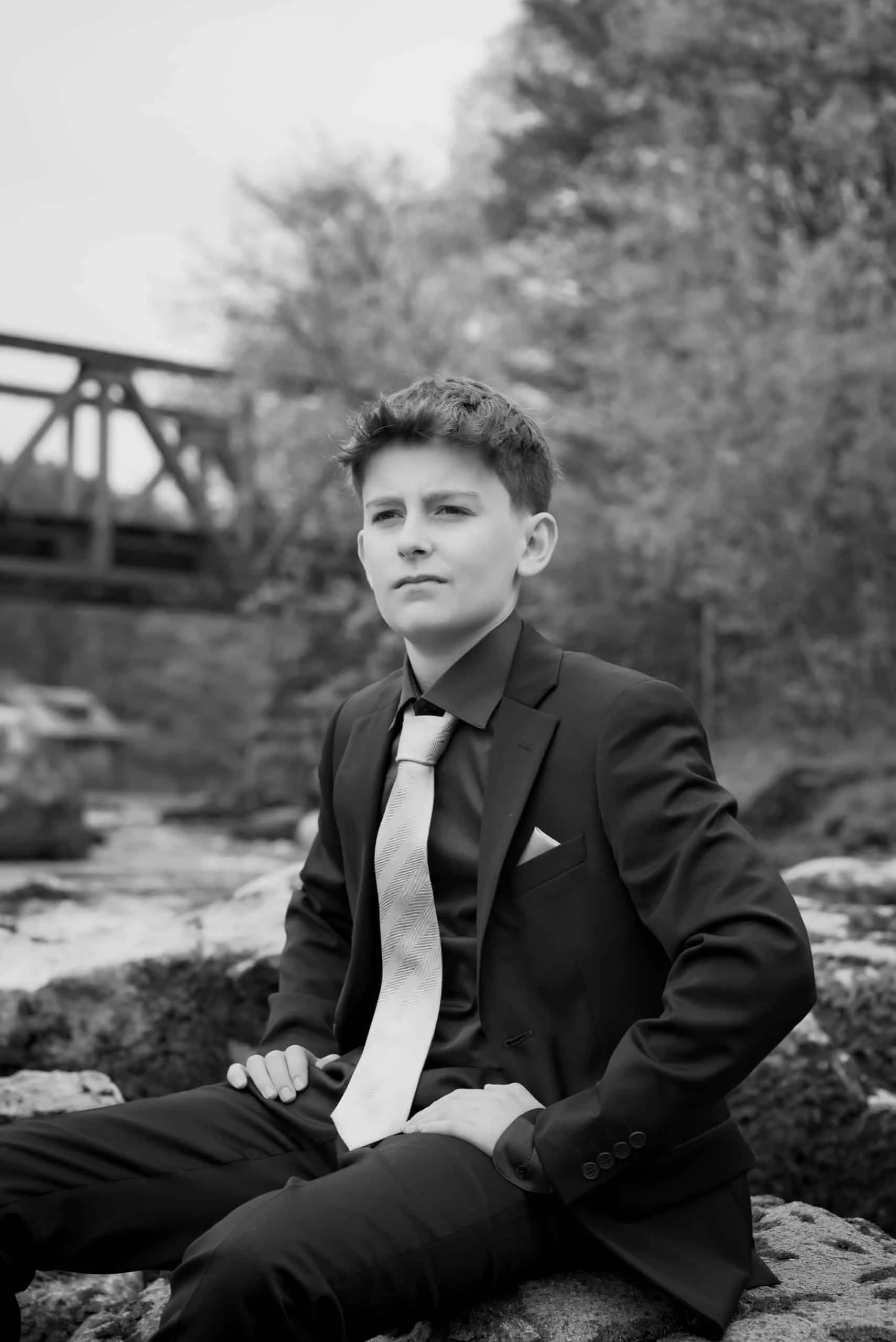 En ung gutt i sort dress og lyst slips sitter på en stein ved en elv under profesjonell portrettfotografering utendørs.