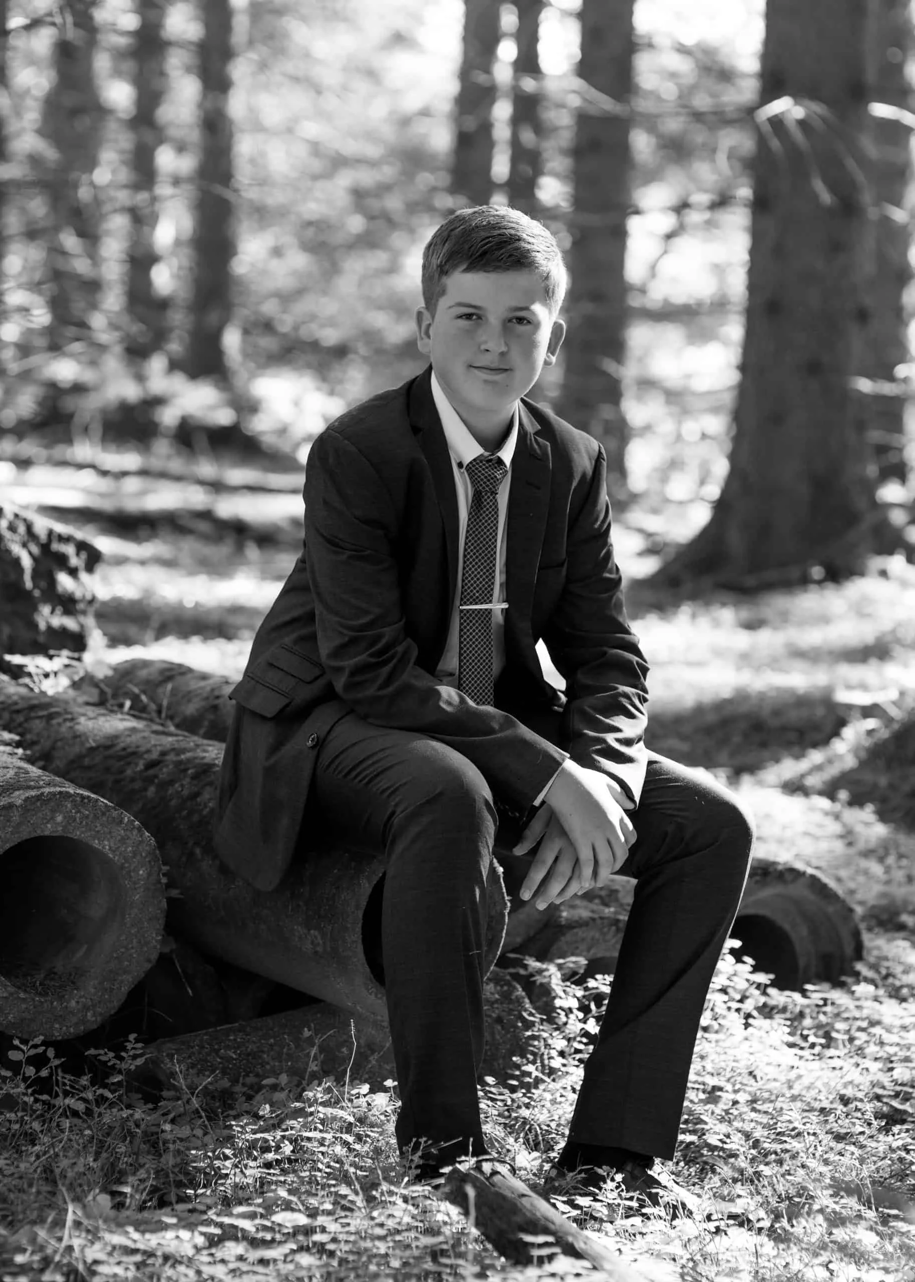 Ung gutt i dress med slips sitter på trestammer i en skog under utendørs familie- og portrettfotografering hos Evig Minne.