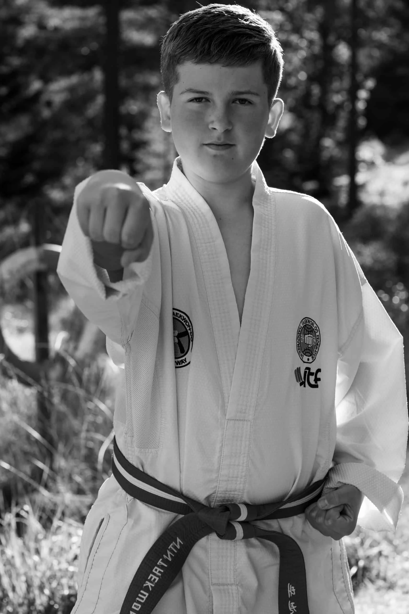 Gutt kledd i hvit taekwondo-drakt med farget belte, utfører et kontrollert slag mot kameraet i profesjonell portrettfotografering utendørs.