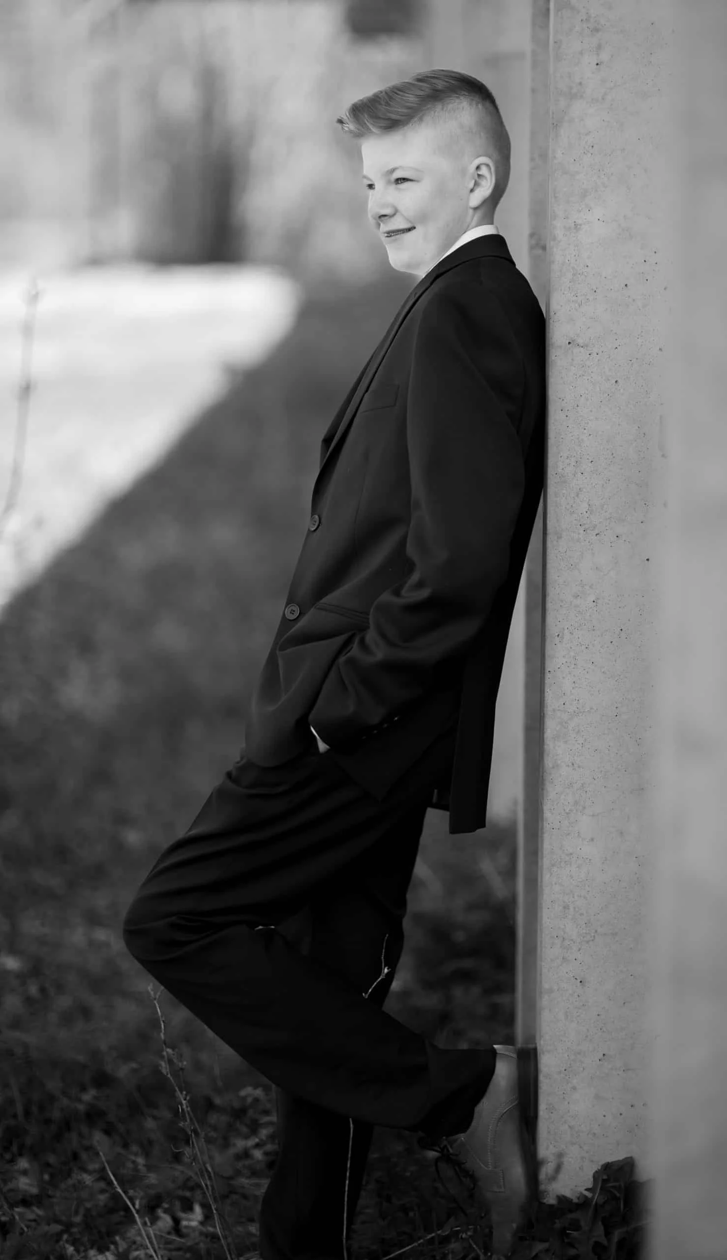 En ung gutt i dress står avslappet lent mot en vegg i sort-hvitt, et elegant portrett fra vår profesjonelle familiefotografering.