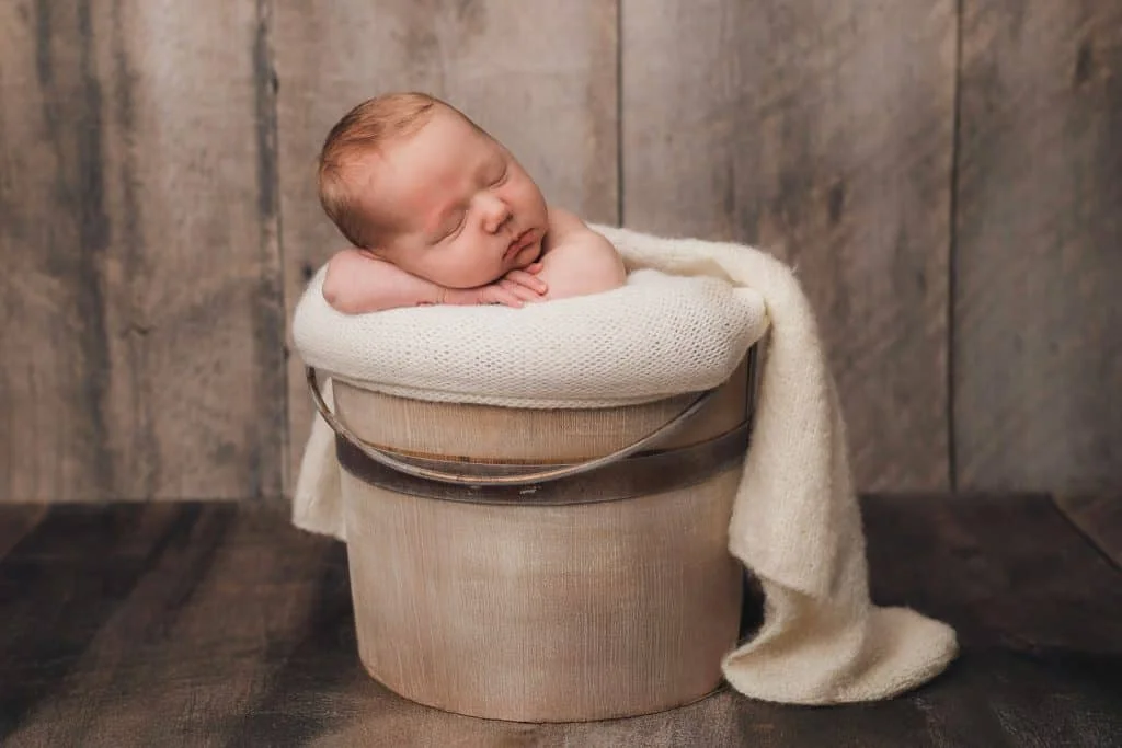 Nyfødt baby sover fredelig med hodet hvilende på hendene i en trebøtte dekket med et mykt, kremhvit teppe i fotostudio.