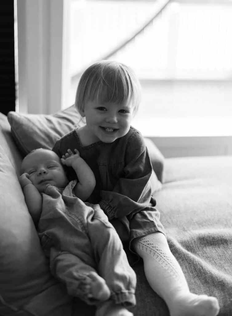 En smilende storesøster holder sin sovende nyfødte bror i et lyst fotostudio under profesjonell nyfødtfotografering.