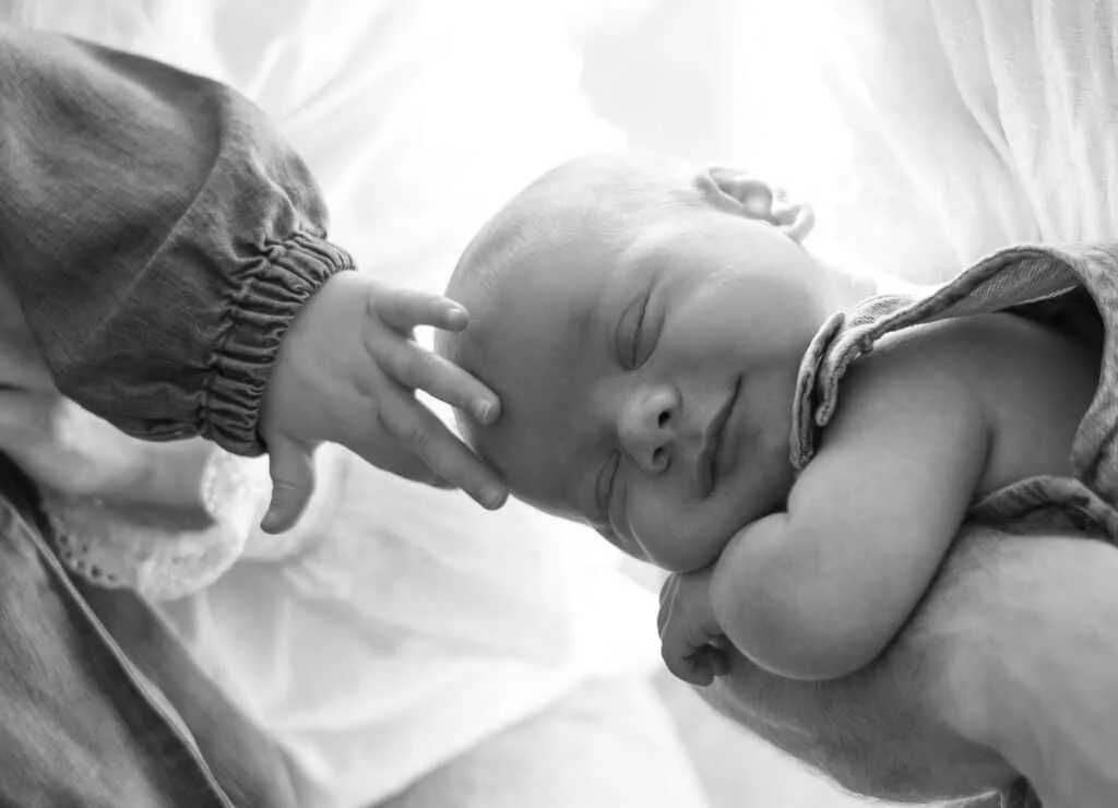 En sovende baby hviler i foreldrenes armer mens et søsken forsiktig berører barnets hode under nyfødtfotografering i vårt lyse fotostudio.