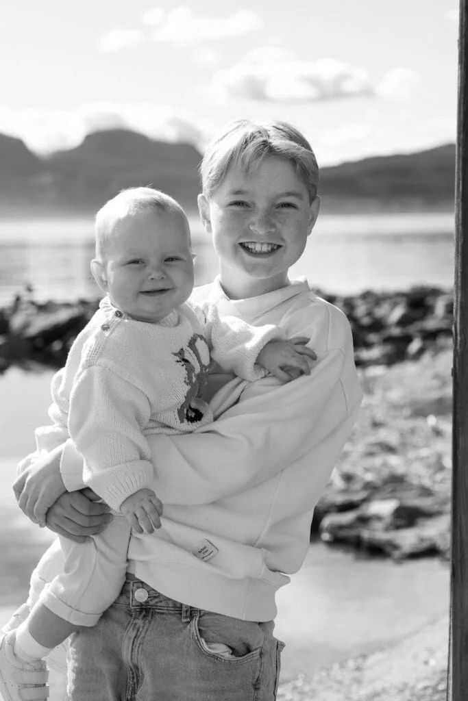 Stolt storesøsken holder en smilende baby i armene utendørs ved sjøen, perfekt for profesjonell babyfotografering og familieportretter.
