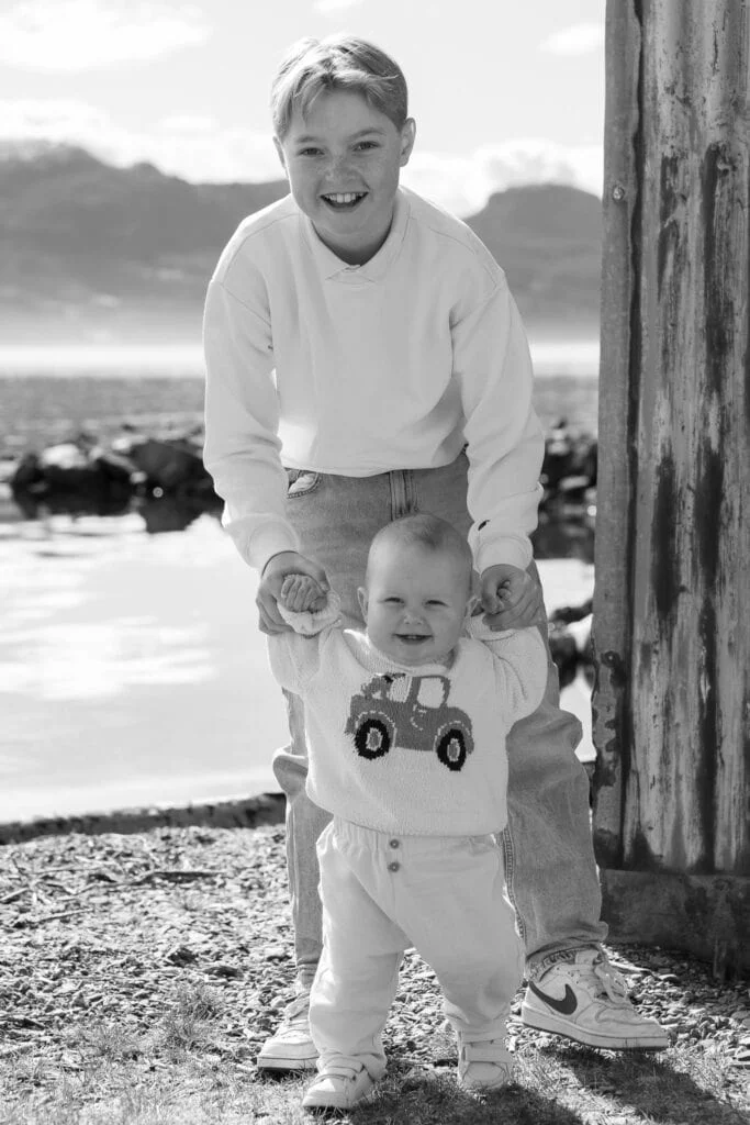 Stolt eldre bror holder en smilende baby i hendene mens babyen tar sine første skritt utendørs ved sjøen.