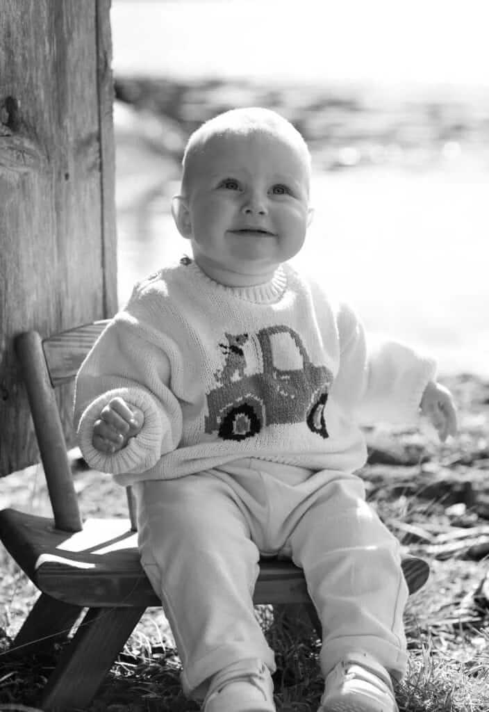 En glad baby sitter på en liten trestol utendørs, iført en strikkegenser med bilmotiv under profesjonell babyfotografering.