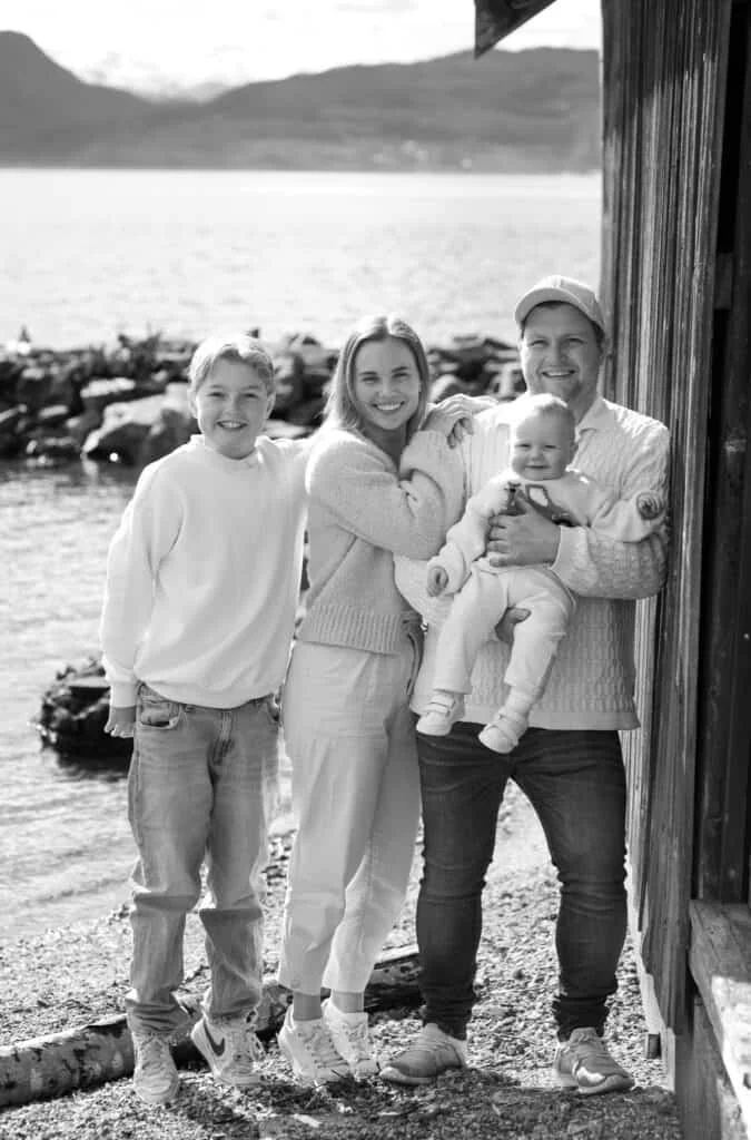 Smilende familie på fire poserer utendørs ved sjøen under en profesjonell familie- og babyfotografering med lys og tidløs stil.