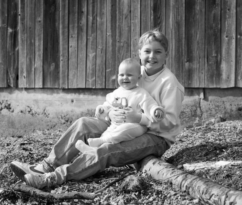 Et eldre barn sitter ute og holder en smilende baby på fanget foran en trevegg, foreviget av en babyfotograf.