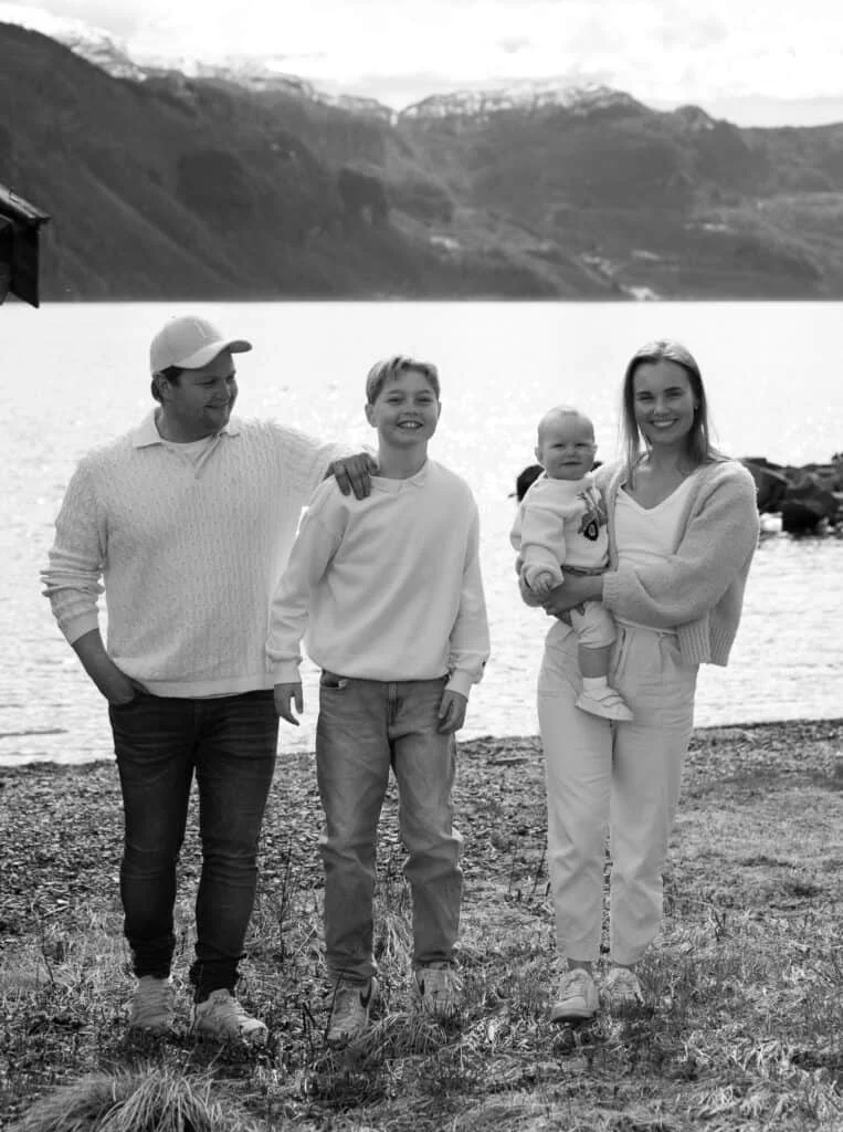 Smilende familie på fire poserer ved vannet under en profesjonell utendørs familie- og nyfødtfotografering med vakker fjellutsikt i bakgrunnen.