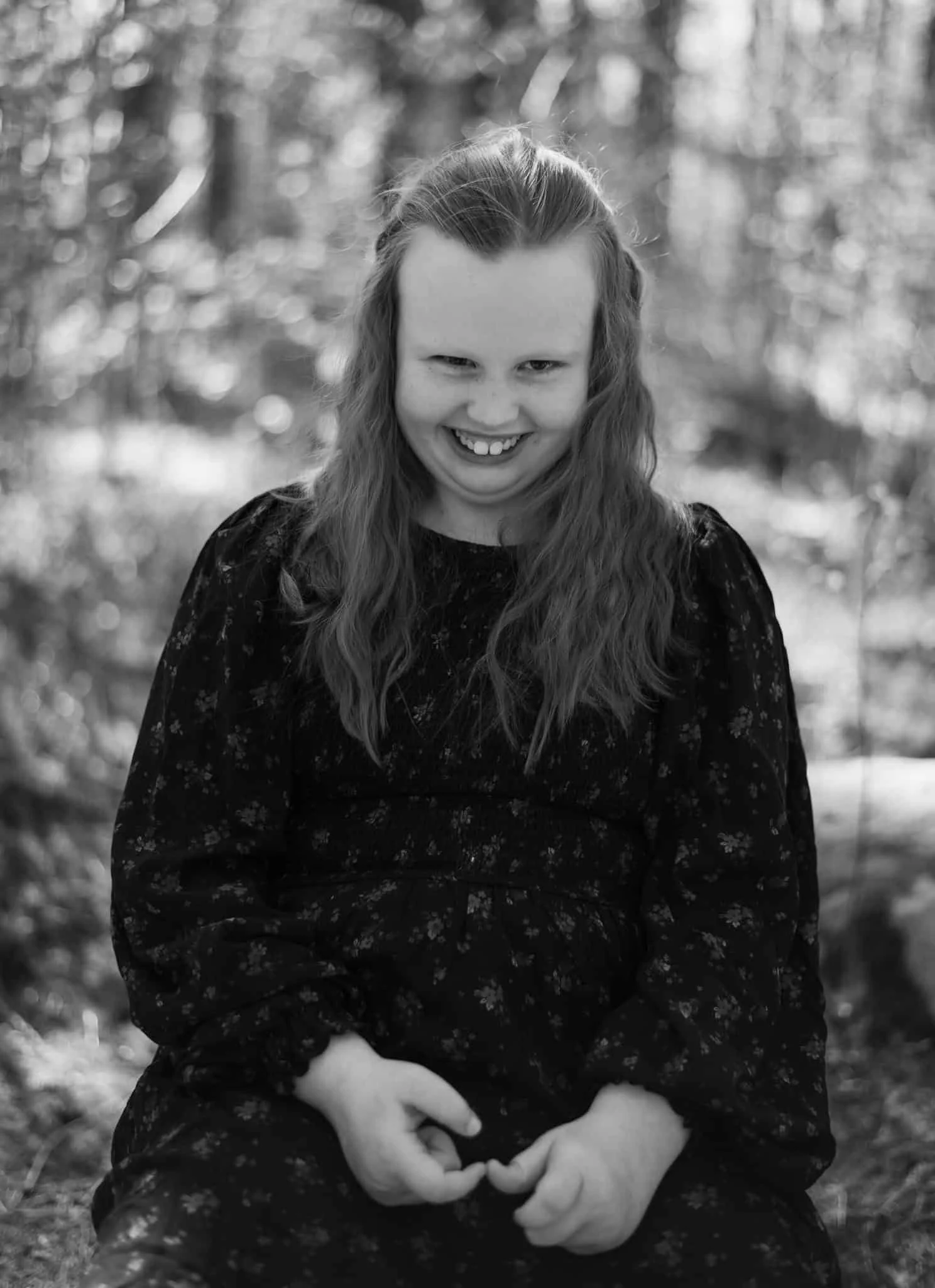 En smilende ung jente i mørk kjole med blomstermønster sitter utendørs i en profesjonell portrettfotografering med duse bakgrunnsdetaljer.