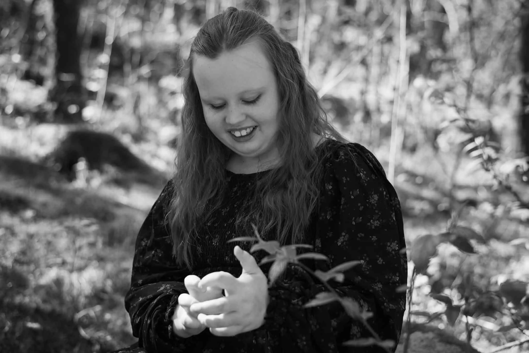 En ung kvinne med langt, bølgete hår smiler ned mot hendene sine i en stemningsfull sort-hvitt utendørsfotografering.