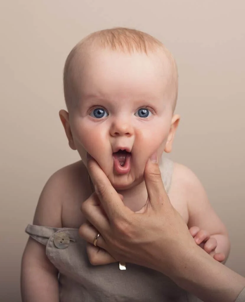 Nærbilde av en baby med blå øyne som får kinnene holdt av foreldrehender under profesjonell babyfotografering i et lyst fotostudio.