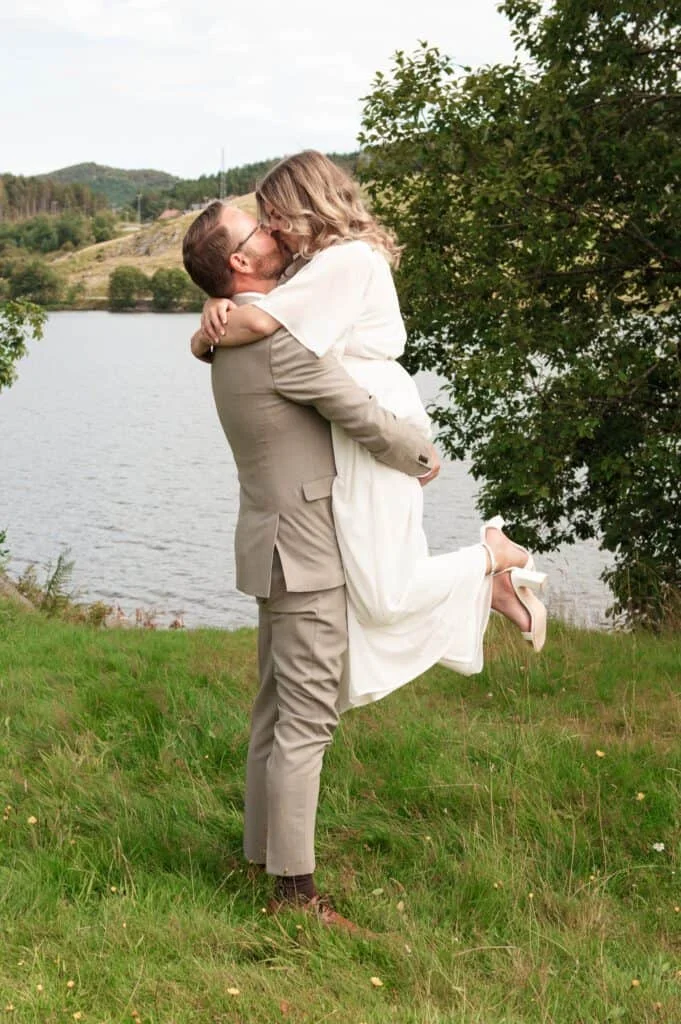 En brudgom i beige dress løfter bruden i hvit kjole utendørs ved en innsjø for profesjonell bryllups- og familie-fotografering.