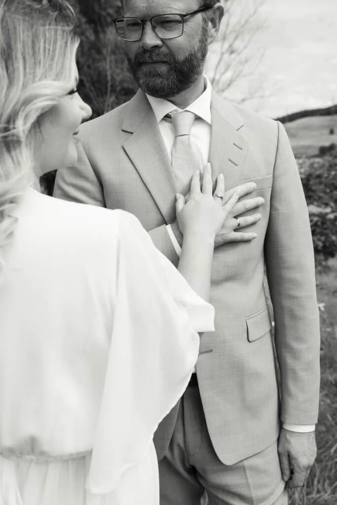 Kvinne med lys kjole legger hånden kjærlig på brystet til en mann i dress i en profesjonell utendørs fotografering.