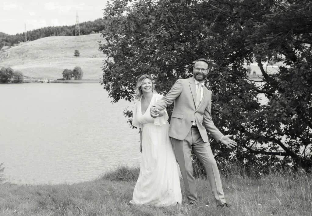 Lykkelig brudepar holder hender og danser utendørs ved en innsjø, et stemningsfullt øyeblikk foreviget av en profesjonell fotograf.