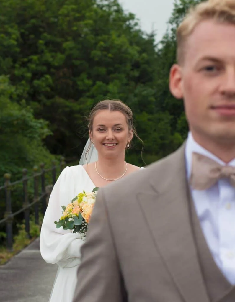 Smilende brud med slør og blomsterbukett i fokus, mens en brudgom i beige dress står uskarp i forgrunnen utendørs.