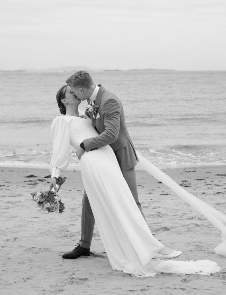 Brudepar i bryllupsklær kysser på stranden, hvor brudgommen bøyer bruden bakover i en romantisk positur under bryllupsfotografering.