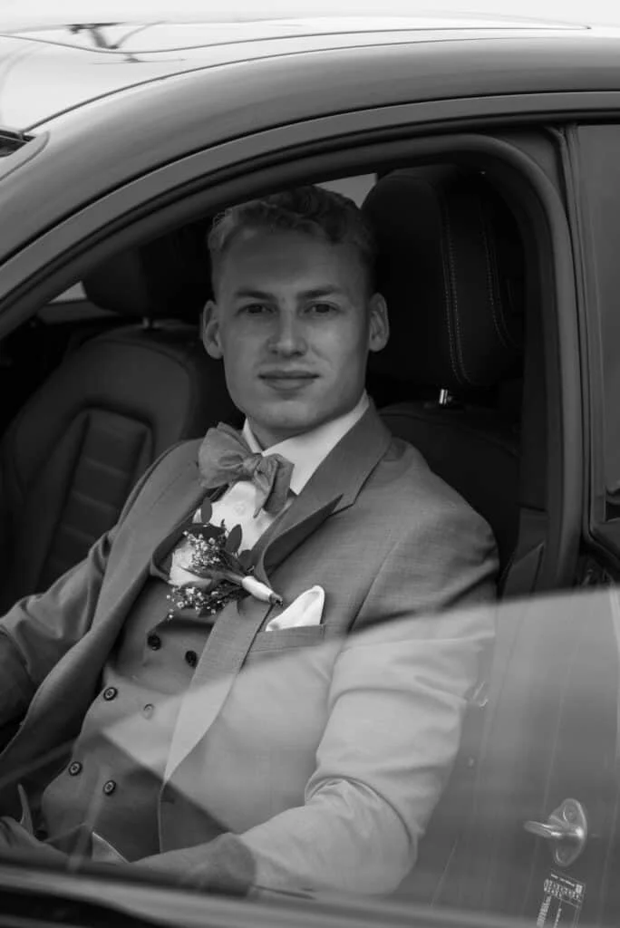 Ung mann i dress med sløyfe og brystblomst sitter i førersetet på en bil, fotografert i profesjonell stil.