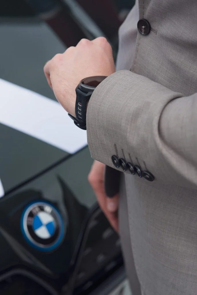 Nærbilde av en person i grå dressjakke med en svart smartklokke, stående ved en mørk bil med BMW-logo.