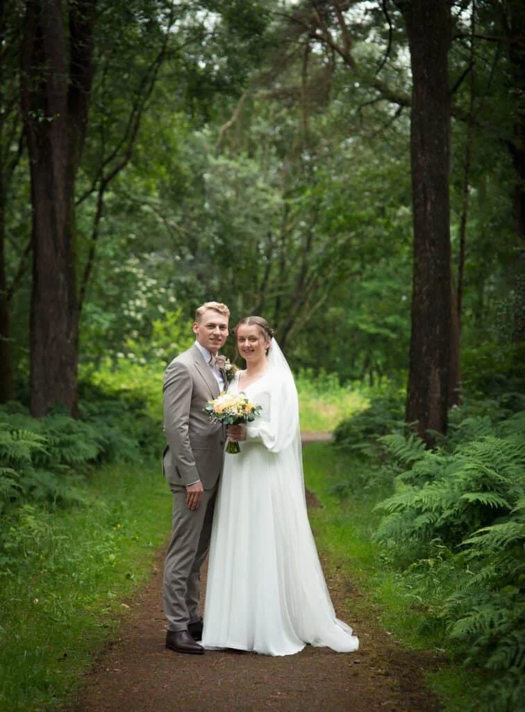 Brudepar står på en skogssti omgitt av bregner, hvor brudgommen i beige dress og bruden i hvit kjole holder blomsterbukett.