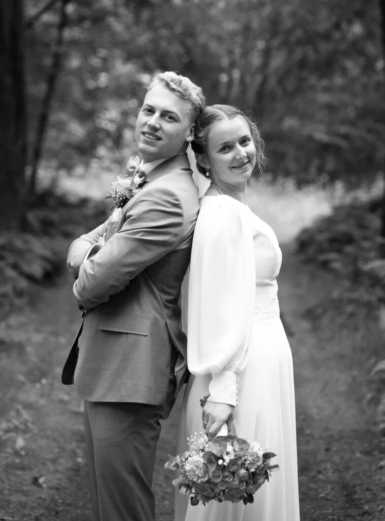 Brudepar står rygg mot rygg i skogen og smiler til kamera under en profesjonell bryllupsfotografering utendørs.