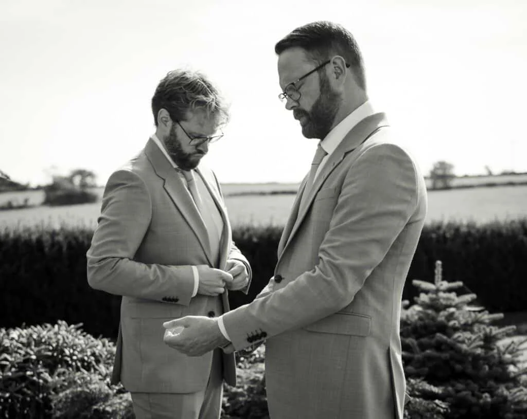 To menn i dress justerer mansjettknapper og jakke utendørs under en profesjonell bryllupsfotografering med fokus på elegante detaljer.
