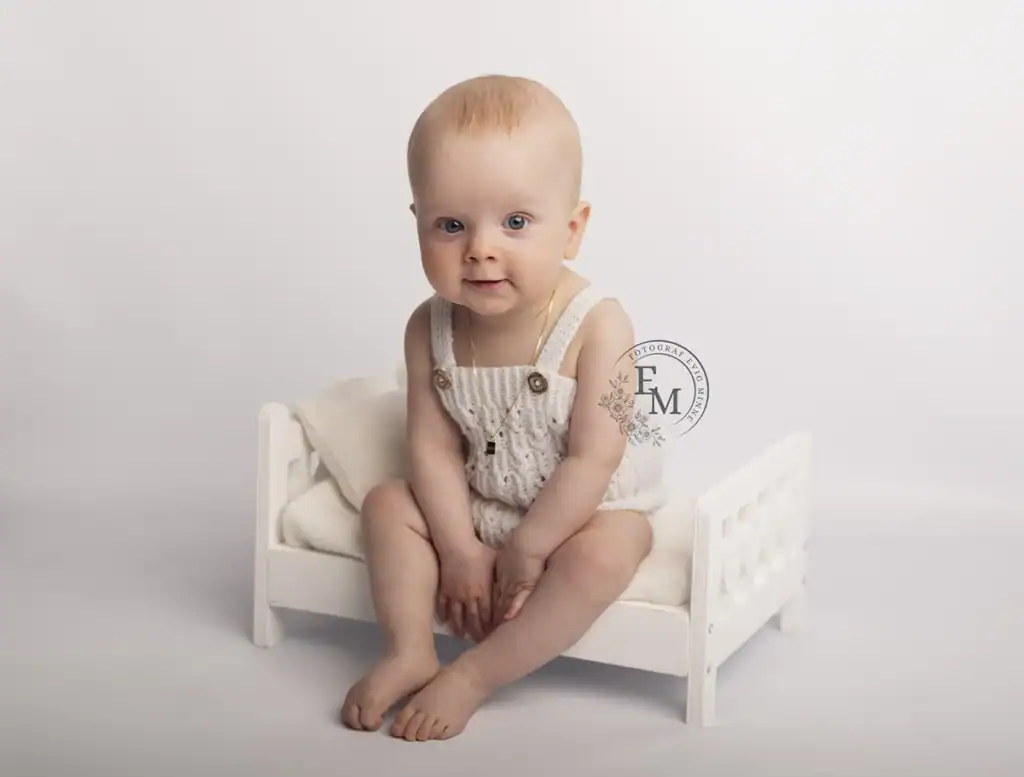 Babyfotografering hos Fotograf Evig Minne av en ettåring i hvit strikka romper som sitter på en liten treseng