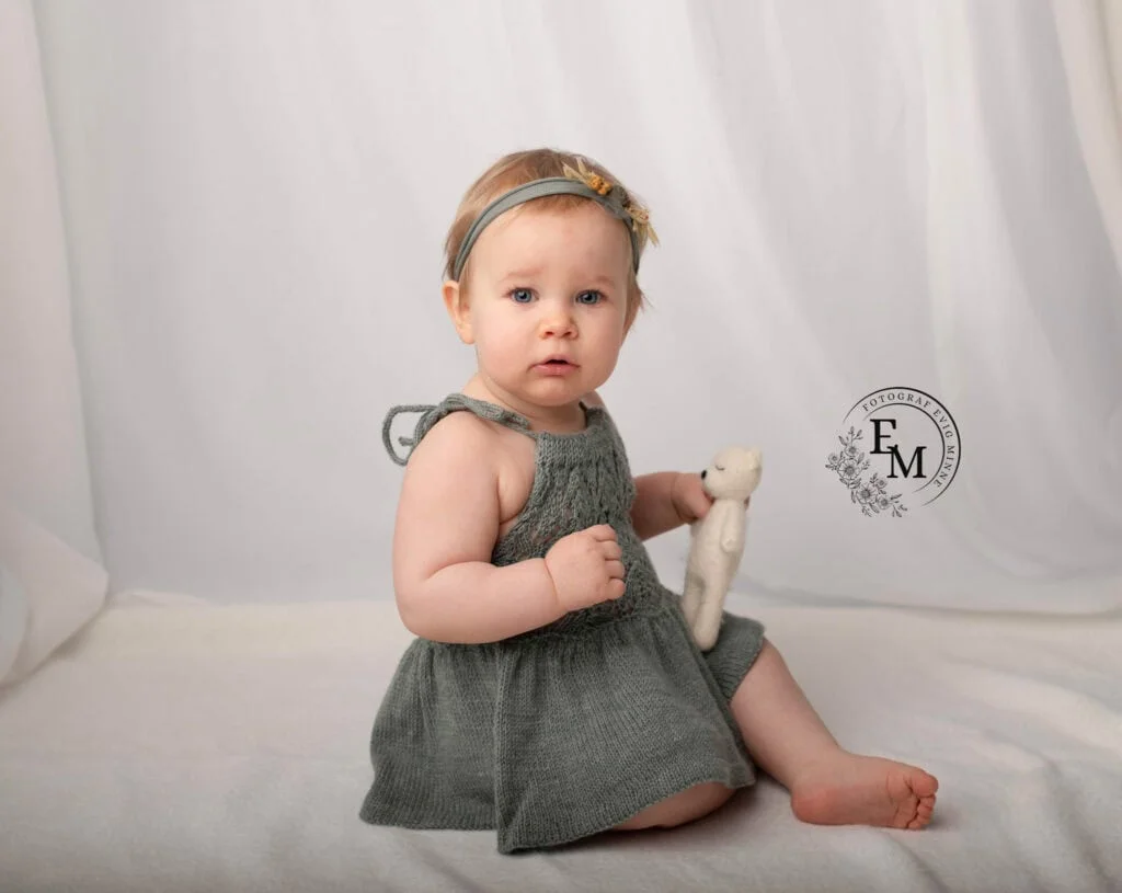 Høykvalitets barnefotografi med babyjente i grå kjole og hodebånd, sittende på hvit bakgrunn.