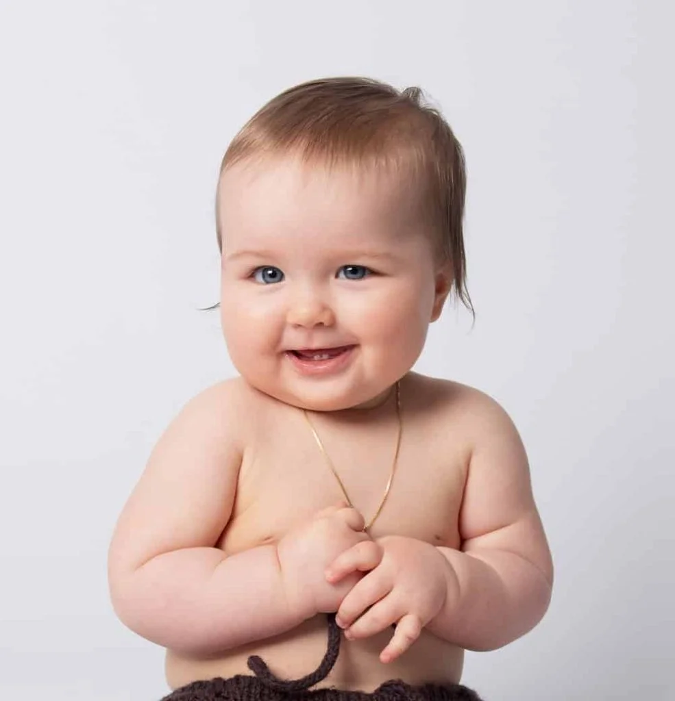 Smilende baby med lyseblå øyne og et tynt gullkjede fotografert i et profesjonelt fotostudio for nyfødtfotografering med lys bakgrunn.