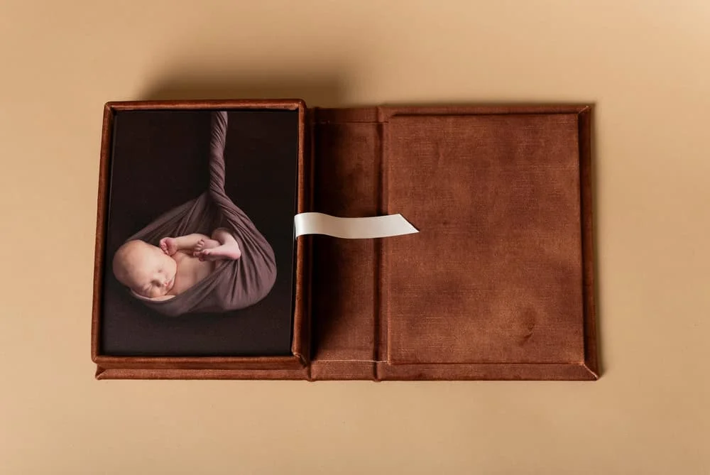 En sovende nyfødt baby svøpt i et brunt stoff, presentert i en elegant brun fotoboks fra vårt nyfødtfotografering-studio.