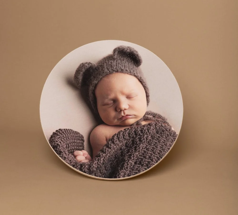 Sovende nyfødt baby med strikket lue med bjørneører, inntullet i et brunt teppe, vist på en rund veggplate i fotostudio.